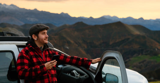 A young man in a plaid jacket standing outside his open car door out in nature among a series of hillls.