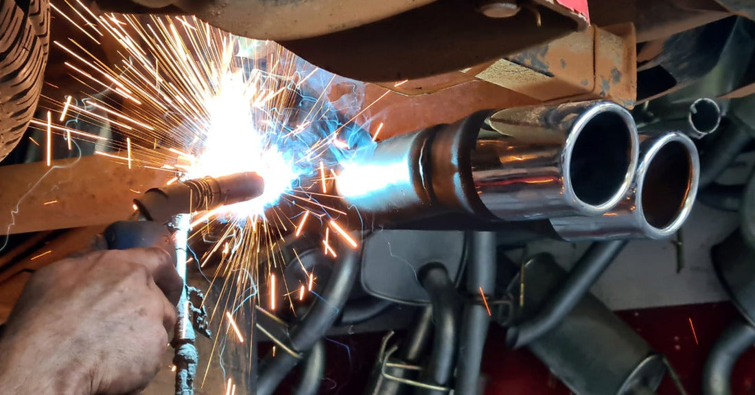 A mechanic using a welding gun to add onto the exhaust pipe that is underneath the car positioned above him.